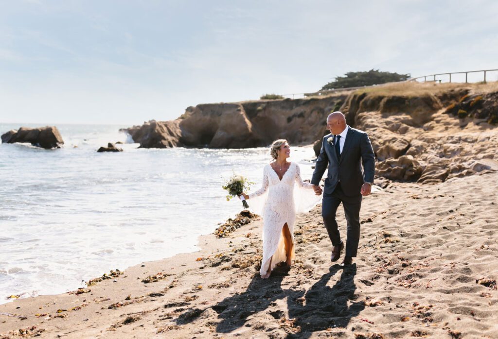 The bride and groom walk together on a beach, surrounded by scenic views and the calming ocean waves.
