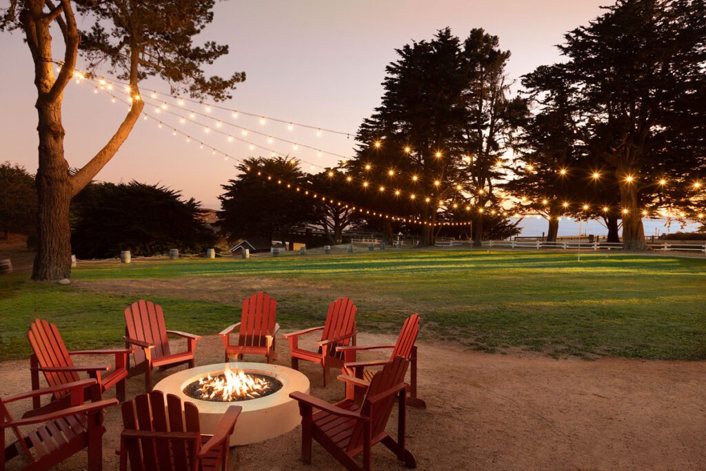 A fire pit with lawn chairs are sat underneath outdoor lights on a wire.